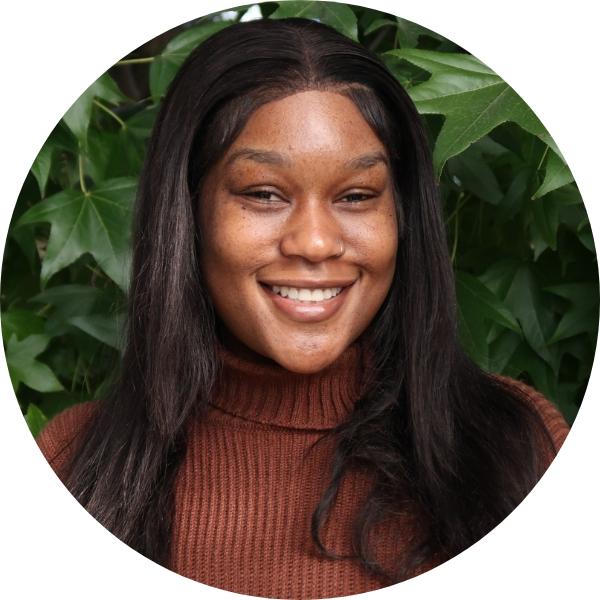 Woman with brown sweater smiling in front of leaves.